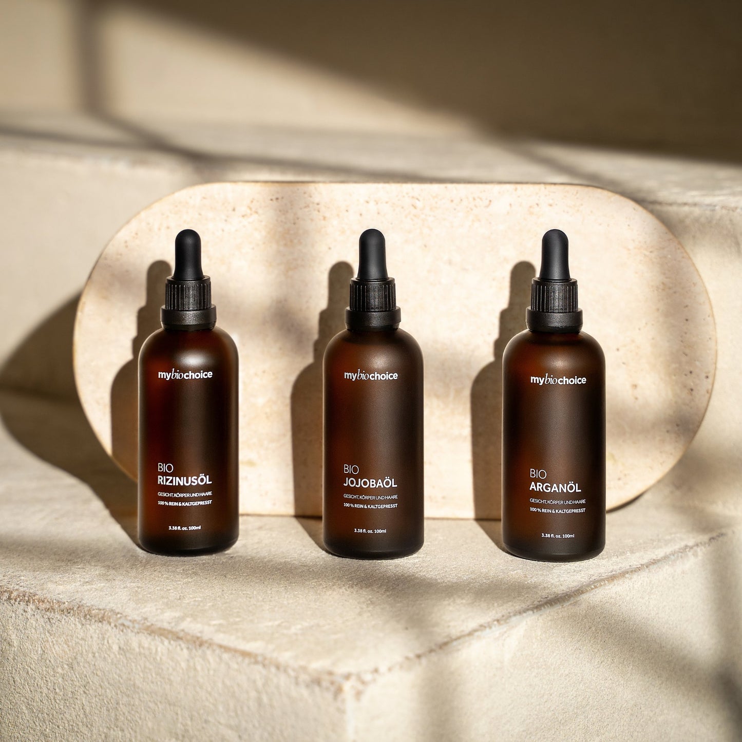 Trzy butelki bio olejków mybiochoice – jojoba, rozmarynowy i arganowy ustawione na jasnej kamiennej powierzchni w naturalnym świetle.