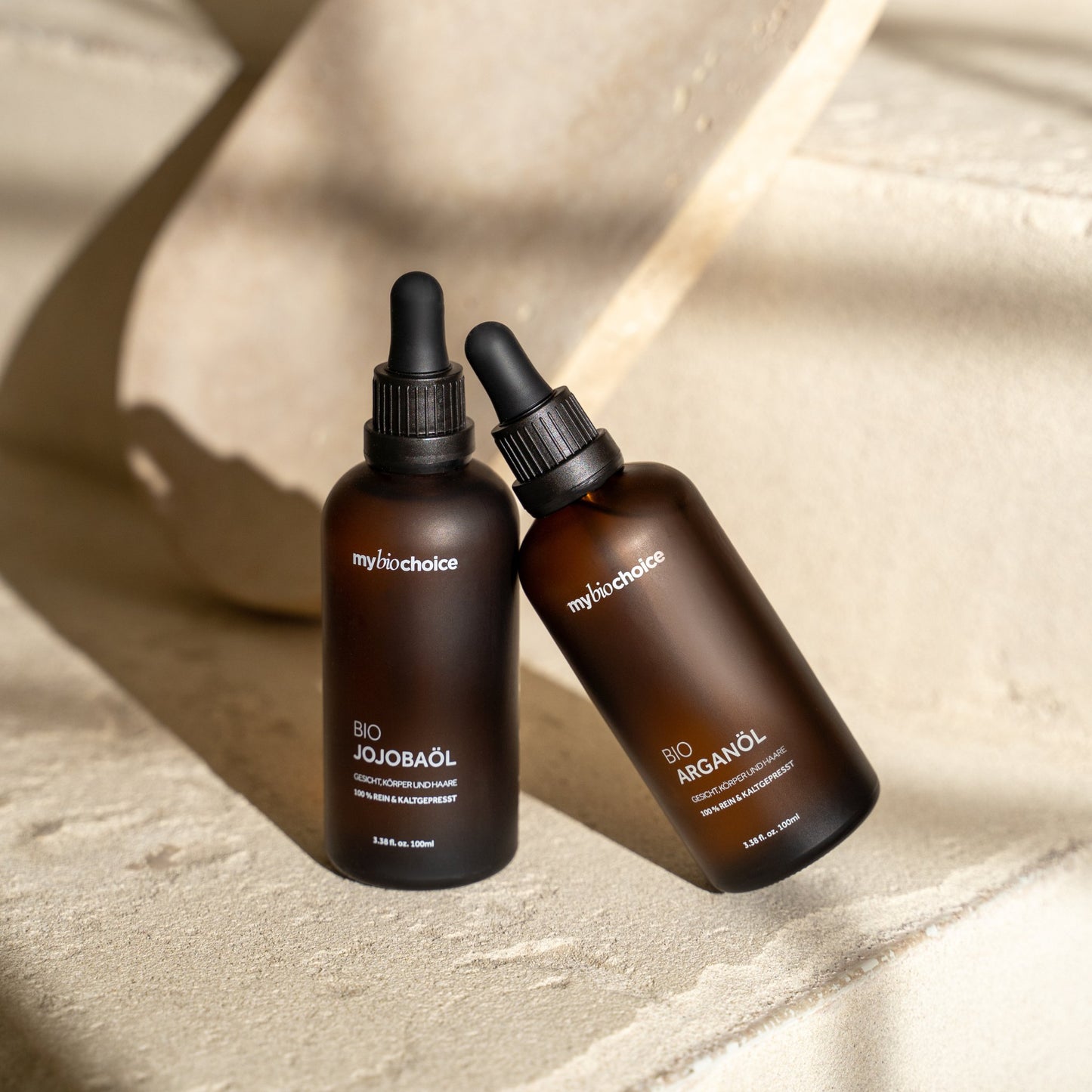 Olejki jojoba i arganowy w brązowych matowych butelkach mybiochoice na jasnej kamiennej powierzchni w naturalnym świetle.