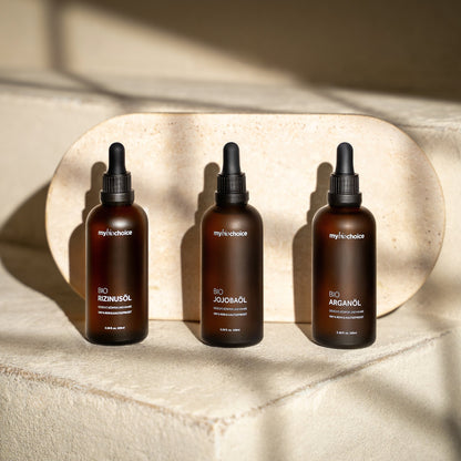Trzy butelki bio olejków mybiochoice – jojoba, rozmarynowy i arganowy ustawione na jasnej kamiennej powierzchni w naturalnym świetle.