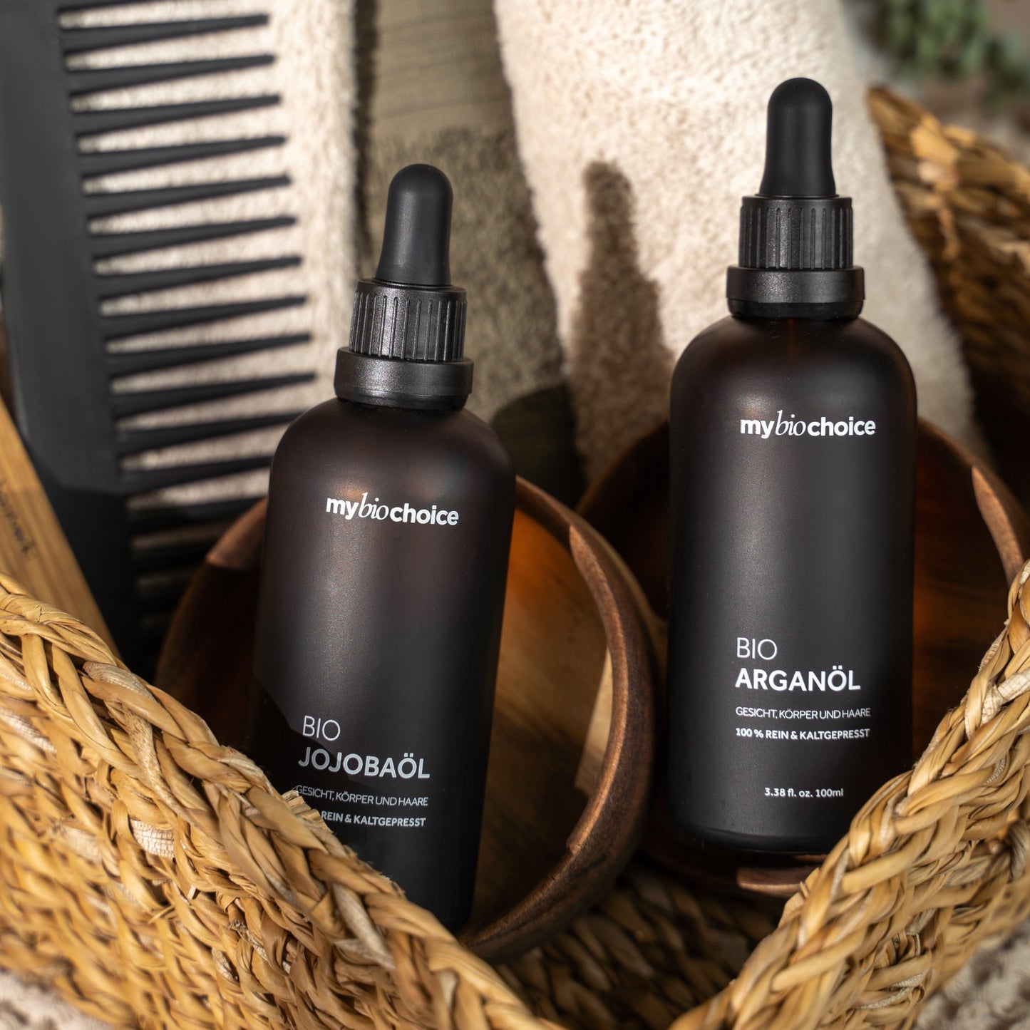 Olej jojoba i arganowy mybiochoice w koszyku z ręcznikami i czarnym grzebieniem – naturalna pielęgnacja włosów.
